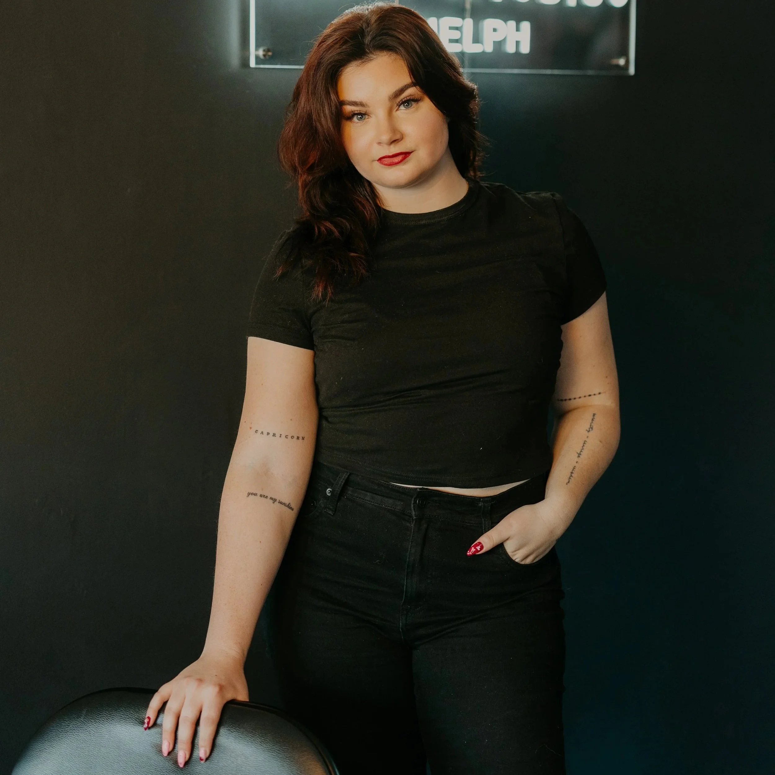 Young woman with brown hair and light makeup wearing a black t-shirt and black pants, standing against a dark wall with a neon sign behind her, holding a chair.