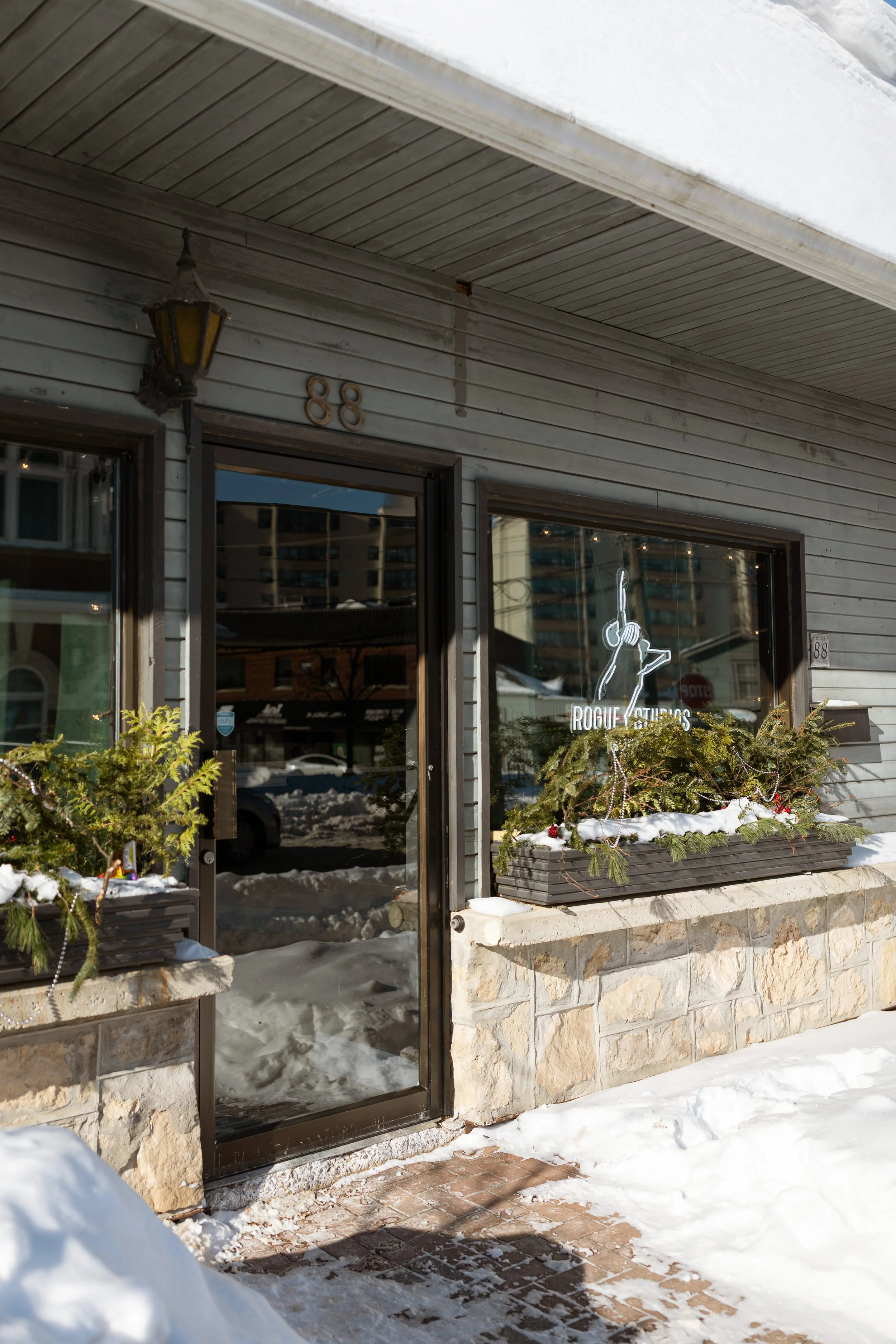 Storefront with stone base, large glass door, the number 88, and a large window with neon sign that reads 'Rogue Studios' featuring a stylized skeleton dancer. Snow covers the sidewalk and window planters.