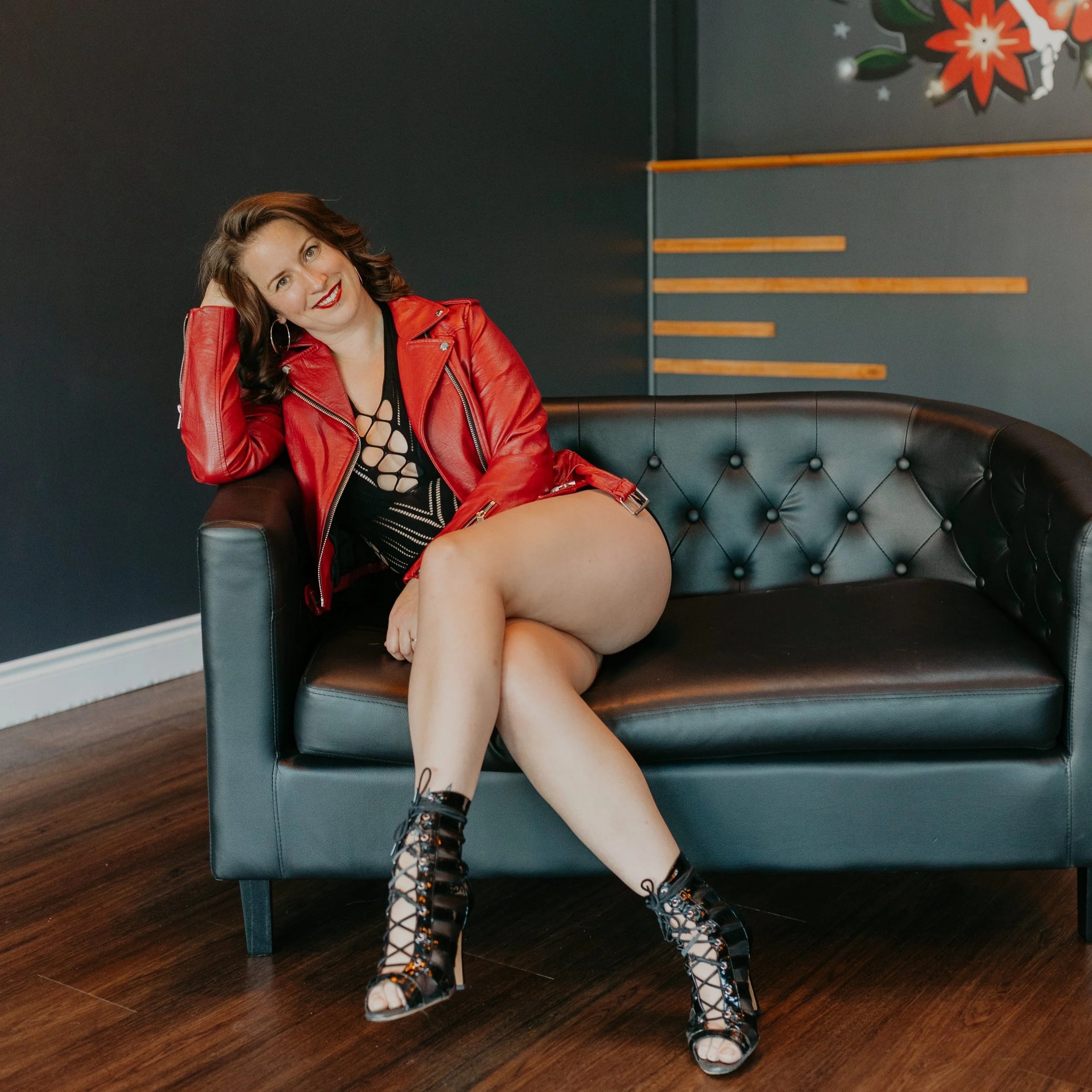 A woman with brown curly hair sitting on a black couch, wearing a red leather jacket and black dance heels, smiling and posing.