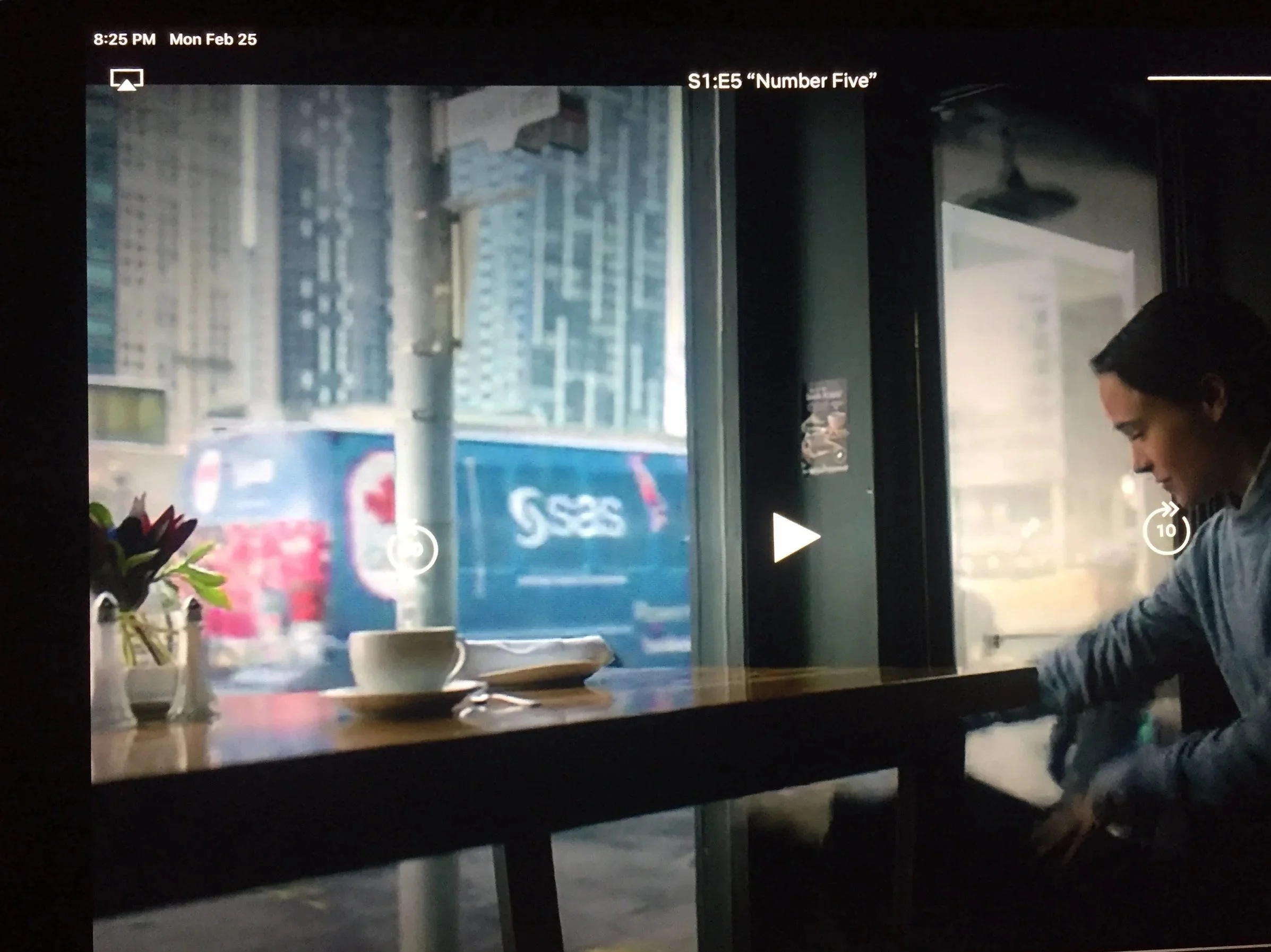 A person sitting by a window during daytime, with a cityscape in the background. Driving by outside is the SAS shuttle for the Canadian Olympics. This is a still from the Netflix series "The Umbrella Academy".