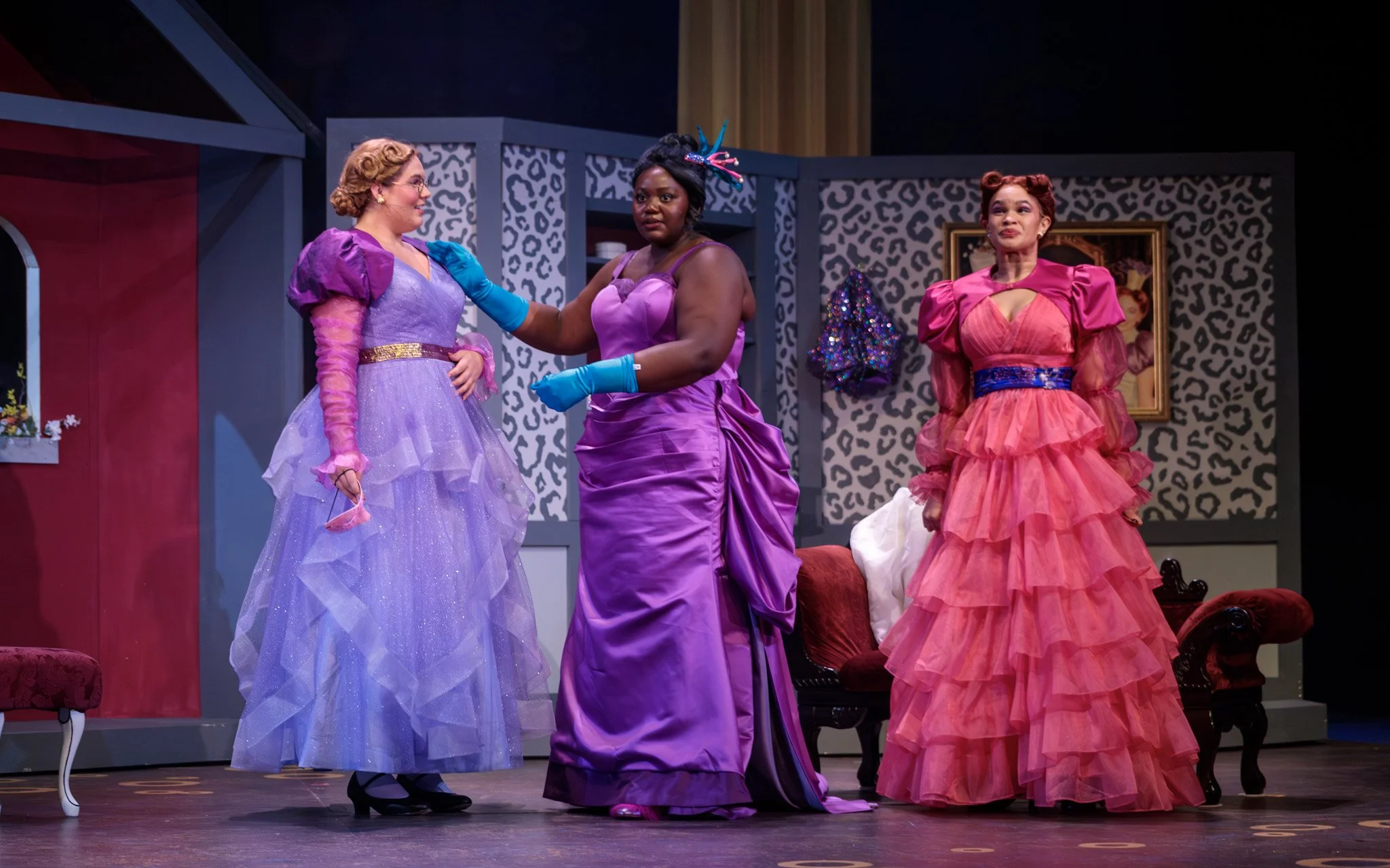 Three women on stage in colorful vintage dresses, one in purple, one in pink, and one in purple satin, appearing to act in a theatrical play with patterned wallpaper and framed pictures in the background.