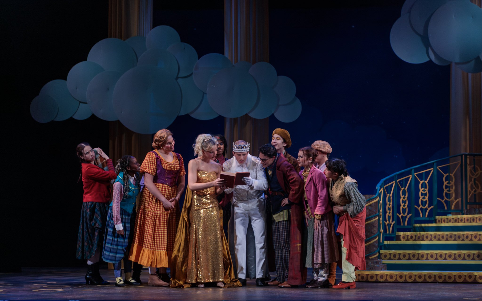 A cast of theatrical performers dressed in colorful costumes gathered around someone reading a book on a stage with large blue balloons and dark backdrop.