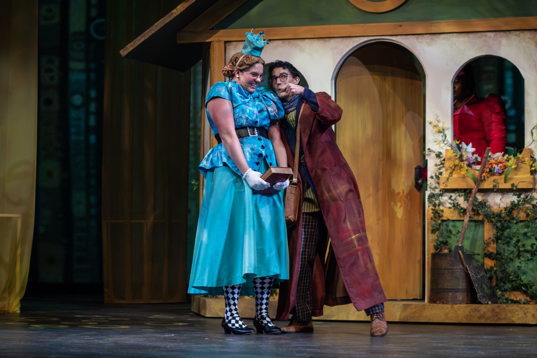 Two women dressed as characters from a theater play, one with a blue dress and hat, and the other with a brown coat, standing on stage with a small house set in the background, smiling and interacting.
