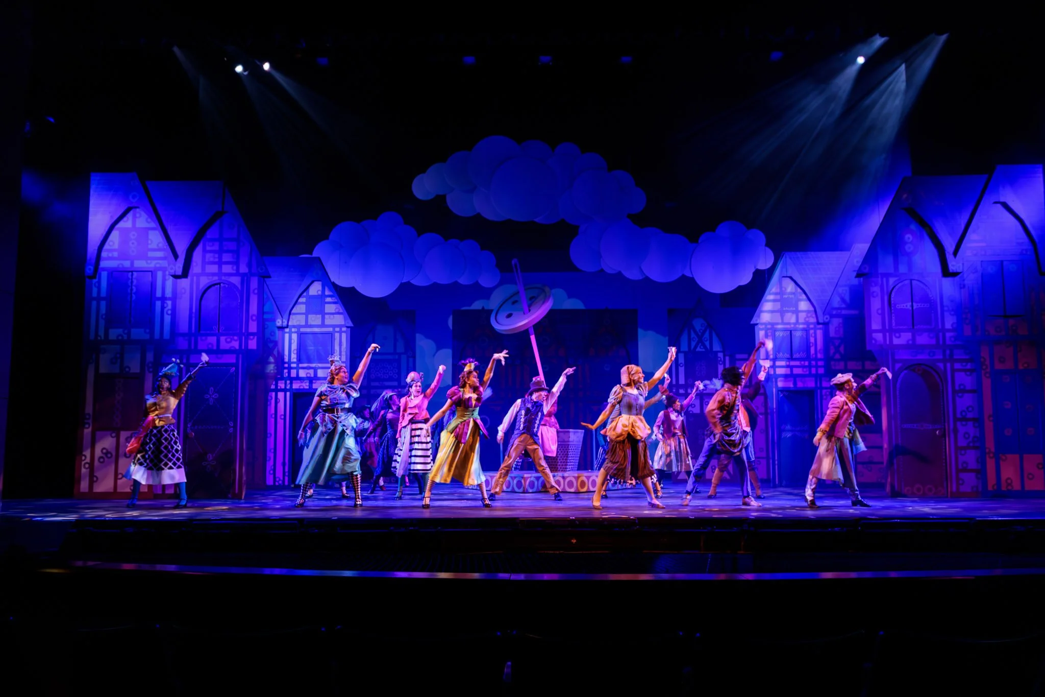 Stage scene from a theatrical musical with performers in colorful costumes, performing a dance in front of a vibrant, cartoon-like backdrop featuring houses, clouds, and a spinning top.