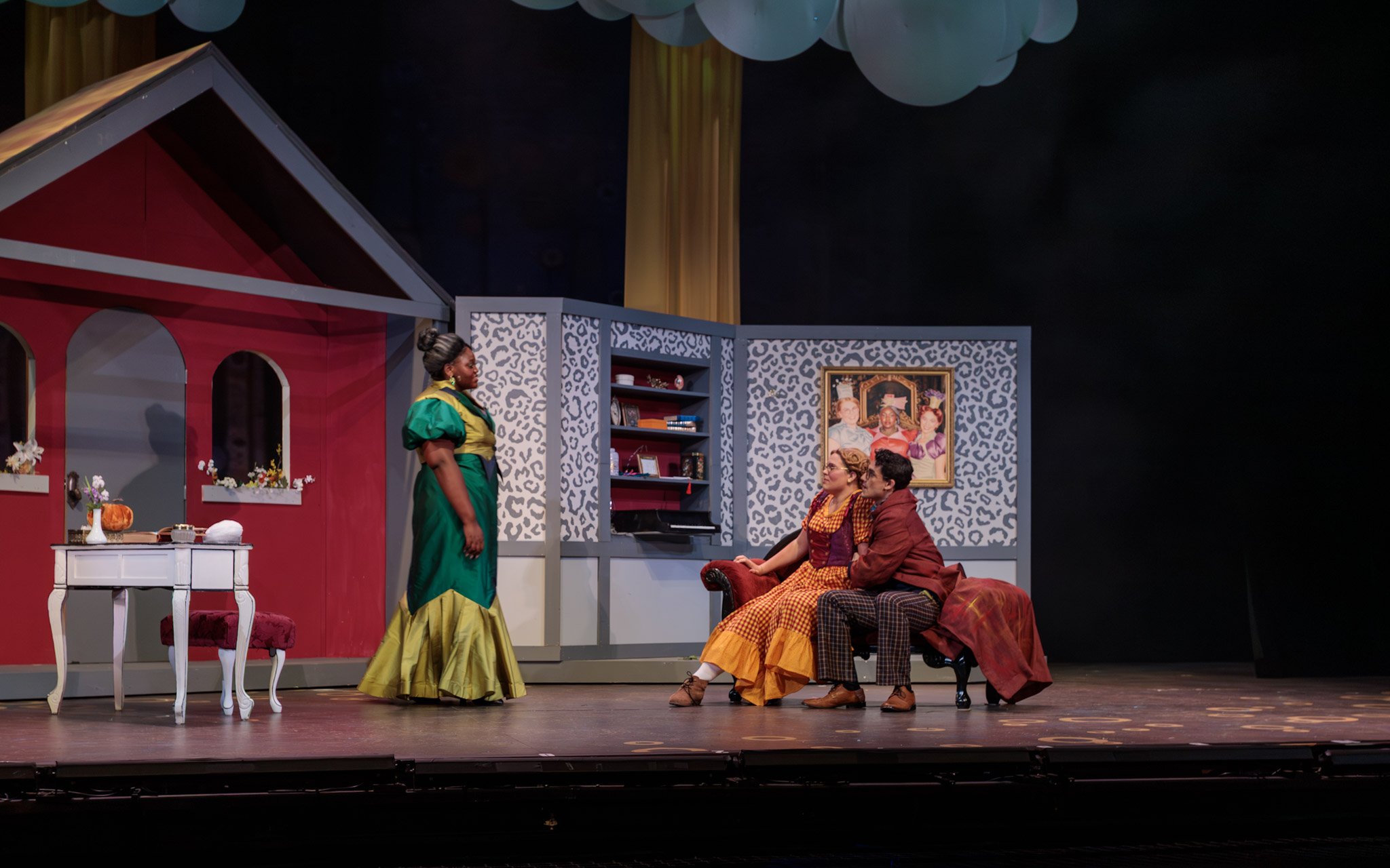Scene from a theatrical play featuring four actors on stage in a room setting with a red and white house facade, a bookshelf, and framed pictures. One woman in a green and yellow dress stands while three others sit on a red and patterned armchair, en