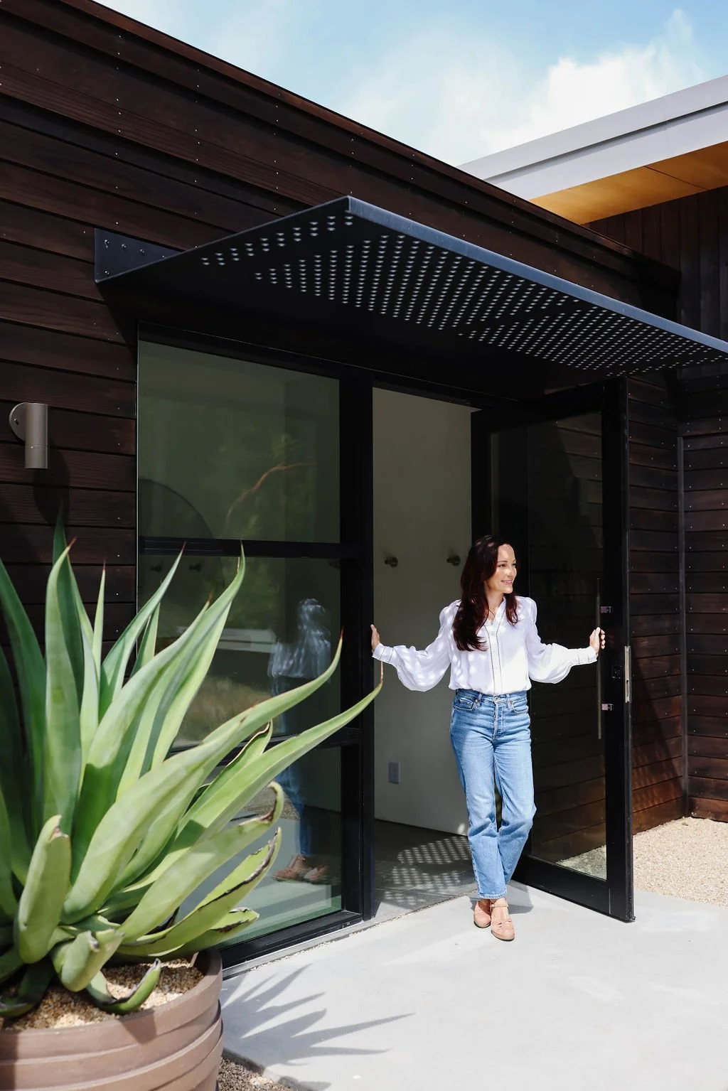 Woman in white blouse and jeans standing at the entrance of a modern house with dark wood exterior, glass door, and large potted plant outside.
