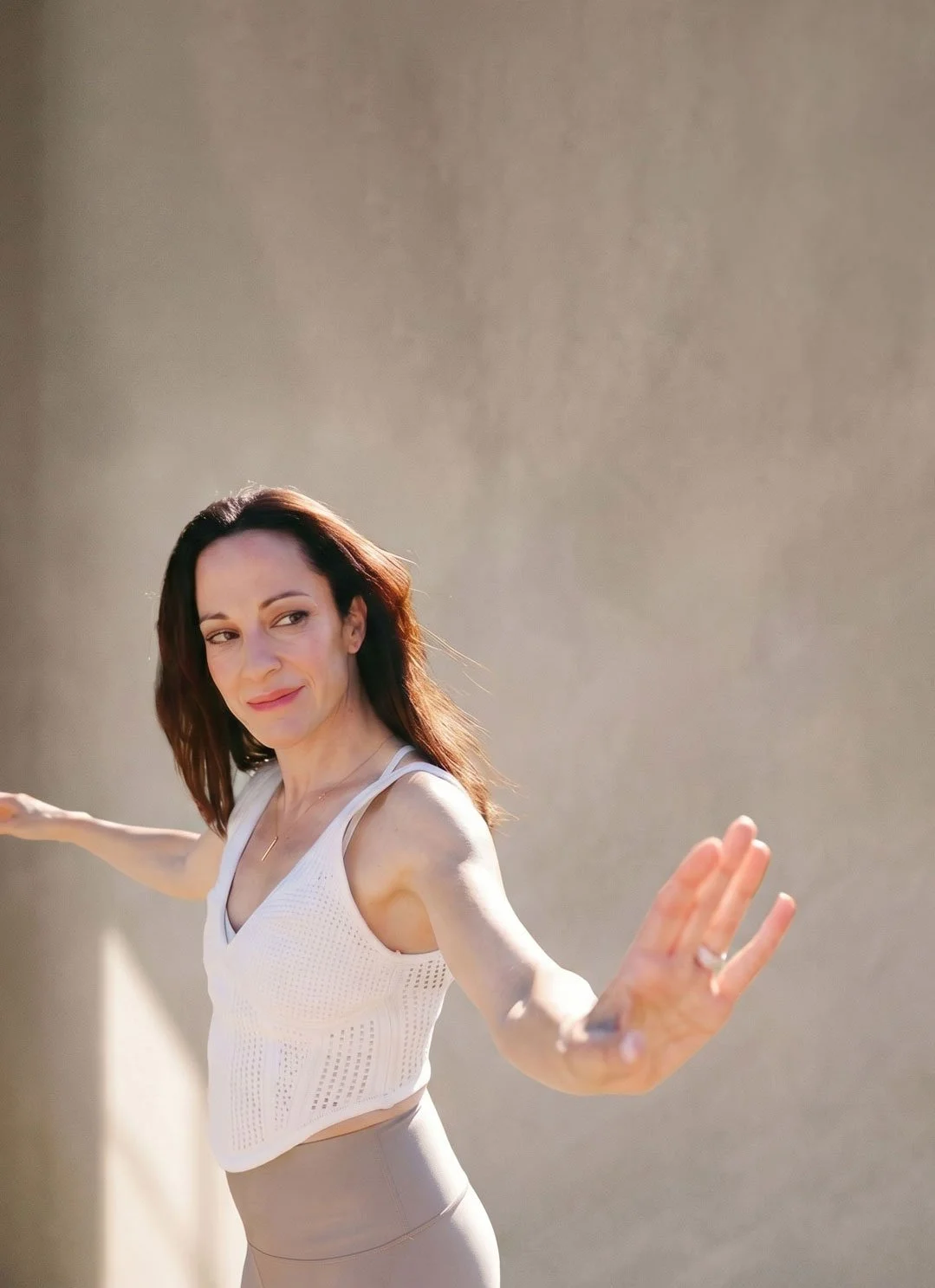 A woman practicing yoga outdoors, leaning against a wall with her arms extended for balance, wearing a white workout top and beige leggings.
