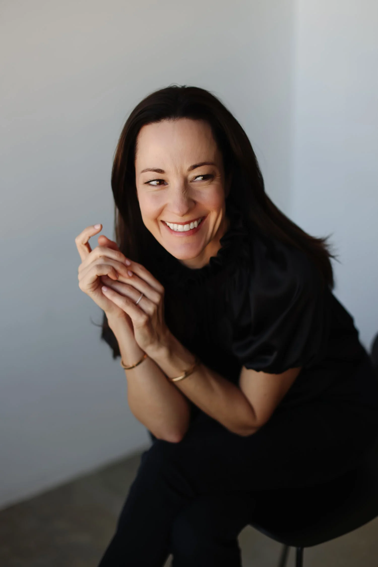 A woman with dark hair, smiling, sitting on a stool in a black top, posing against a neutral background.