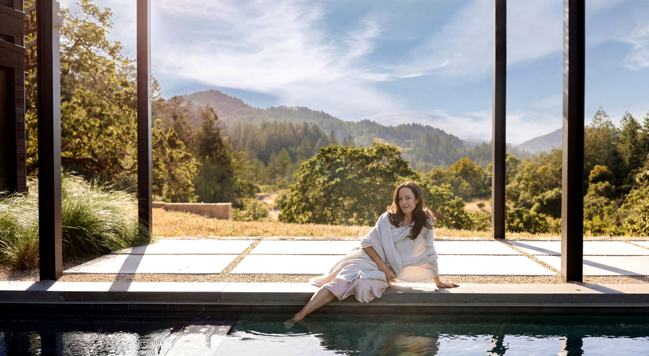 A woman in a white blanket sitting by an outdoor pool with a scenic mountain and forest view in the background.
