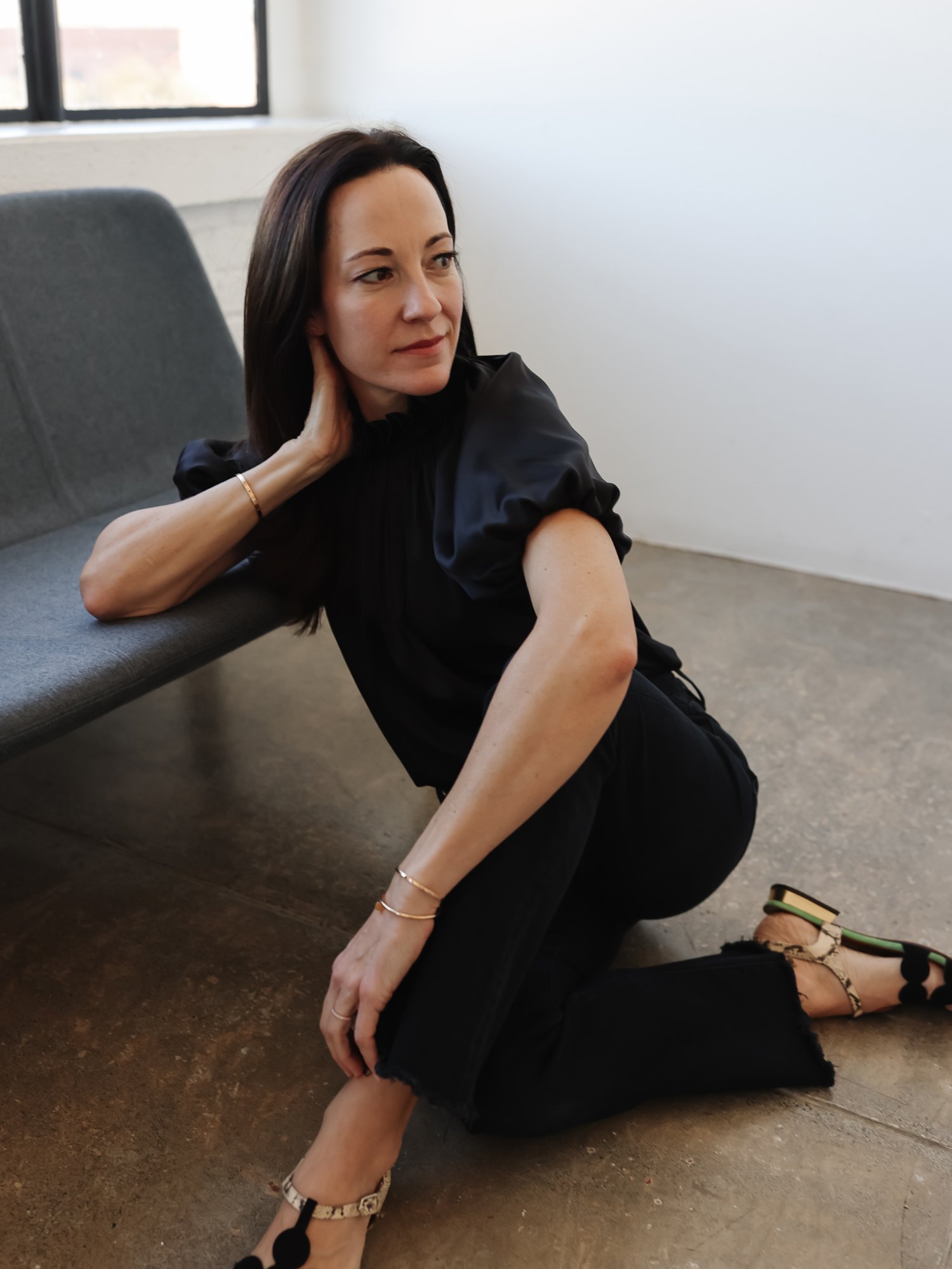 A woman with dark hair, wearing a black top and black pants, is kneeling on the floor indoors next to a gray bench. She is looking off to the side with her hand resting on her neck and the other on her knee. She is wearing pointed high heels with a pattern and green accents.