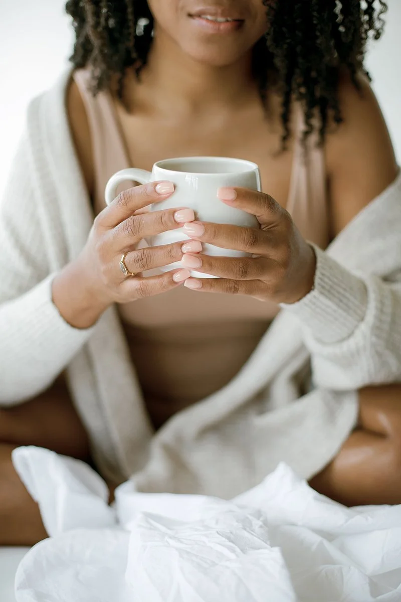 A woman with curly hair holding a white mug and smiling softly, sitting on a bed with white sheets, wearing a beige top and a white cardigan.