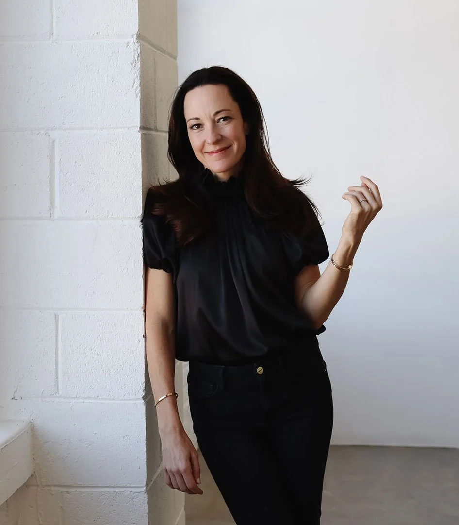 A woman with long dark hair is smiling and leaning against a white wall in a minimalistic room, wearing a black blouse and black pants.