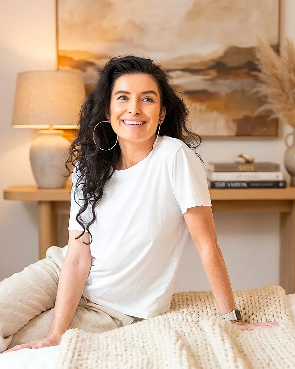 A woman name Marika Jozwiak with dark curly hair smiling, wearing a white t-shirt and large hoop earrings, sitting on a cream-colored knitted blanket in a cozy living room with a lamp, artwork, and a stack of books in the background.