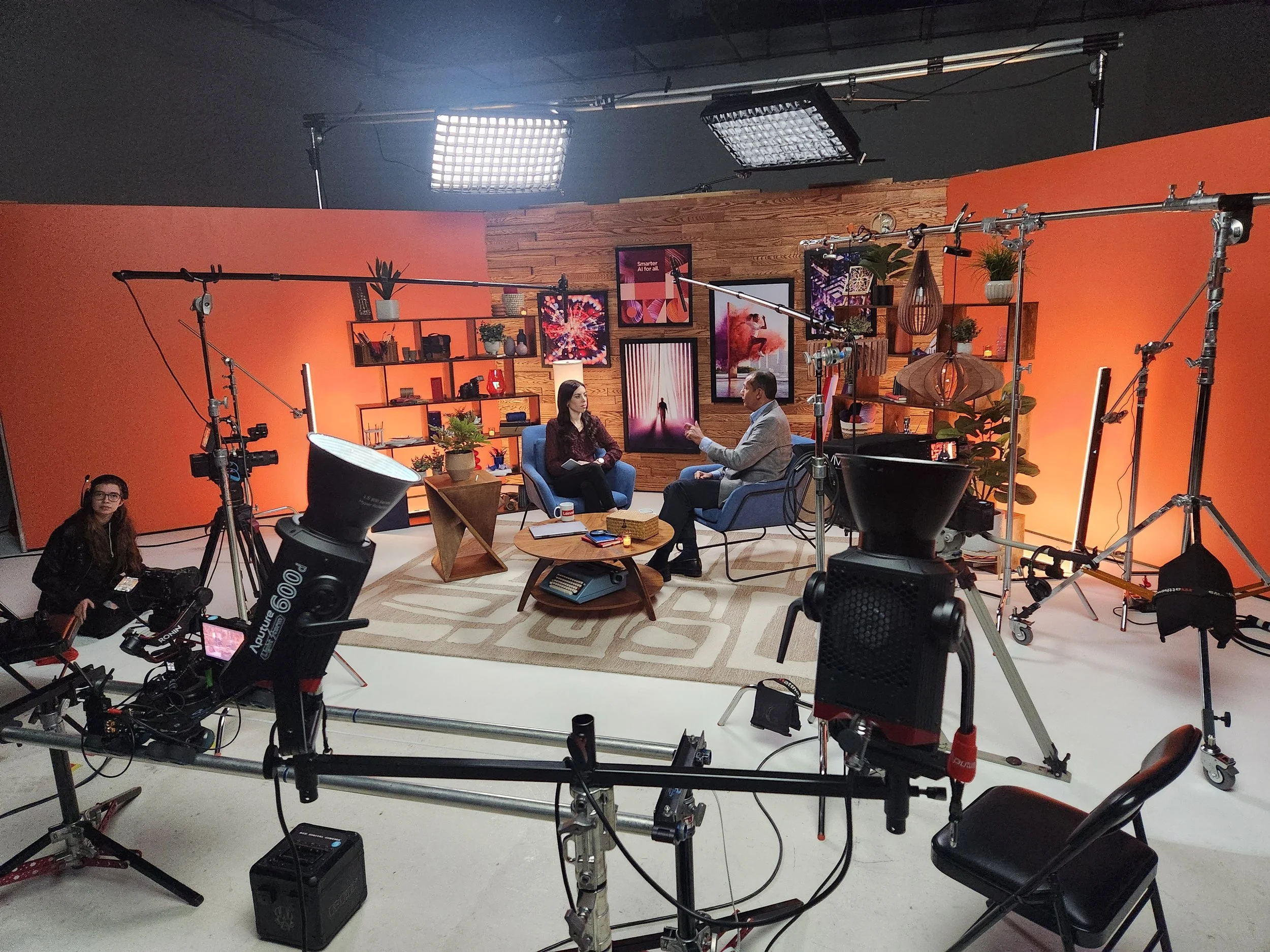 A podcast studio set with two people in conversation surrounded by professional lighting equipment, cameras, and monitors, with a colorful background on the wall.
