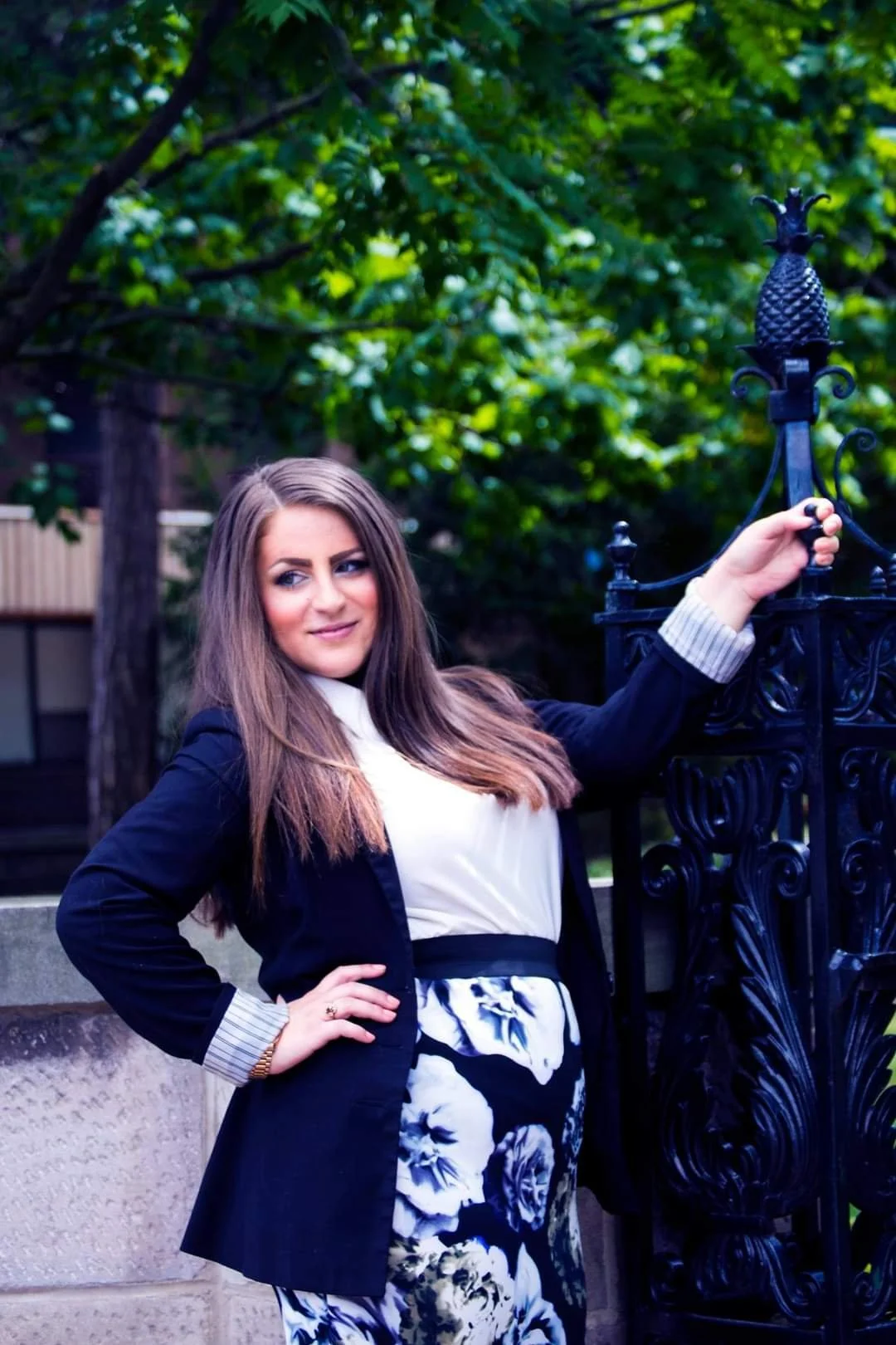 A woman with long brown hair wearing a black blazer with white cuffs, a white top, and a floral skirt standing outdoors near a black ornate gate with green trees in the background.