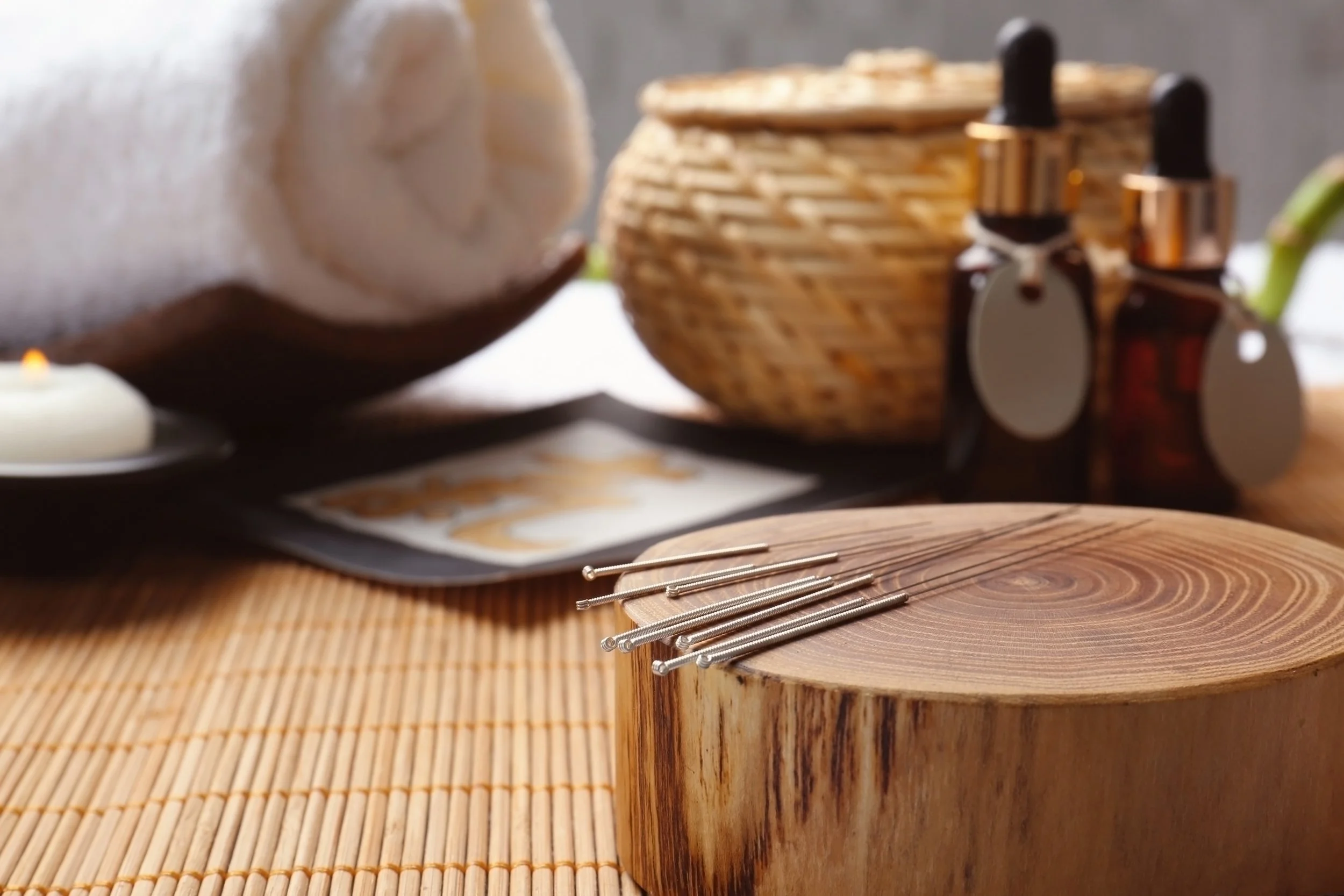 Close up on acupuncture needles, bottles of essential oil, a rolled up white towel and a wicker basket.