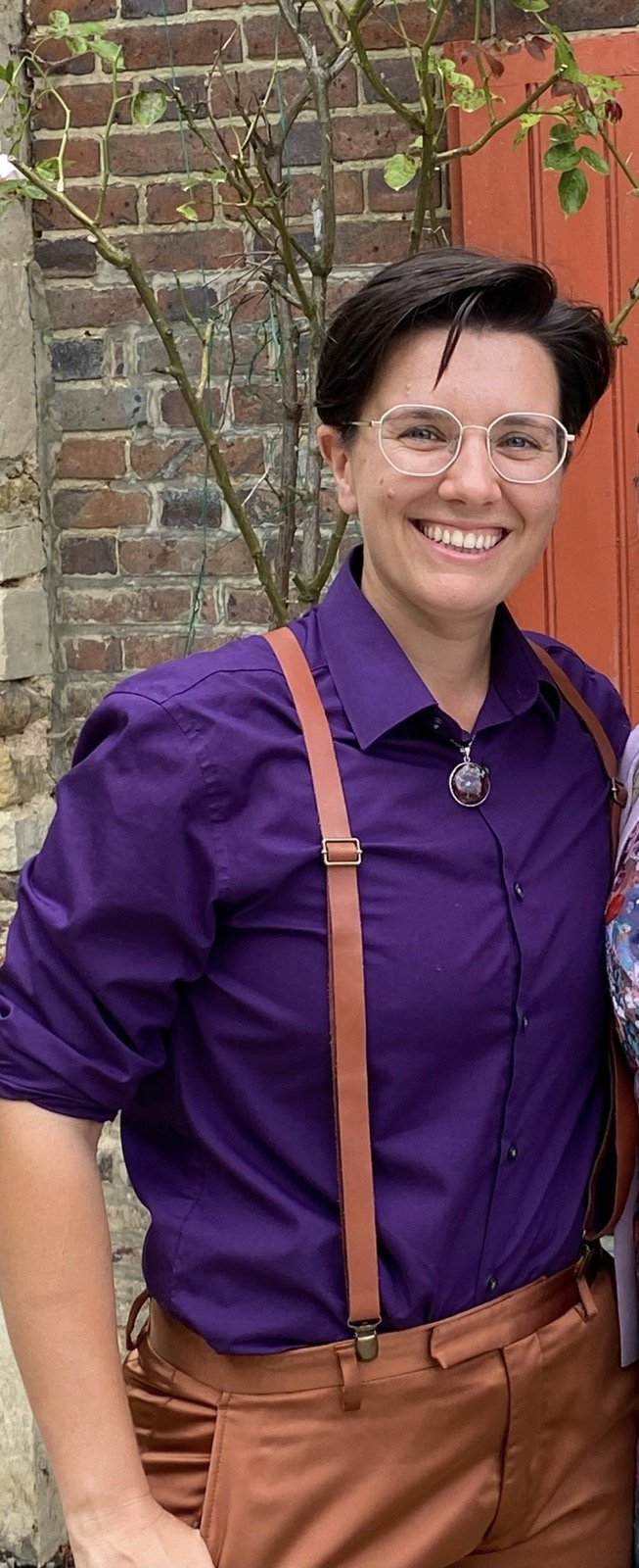 Kyng stands outside in front of a brick wall with a big smile. They are a white skin person with short dark hair wearing glasses, a purple button down shirt, a splash of color with a necklace, and thin suspenders.