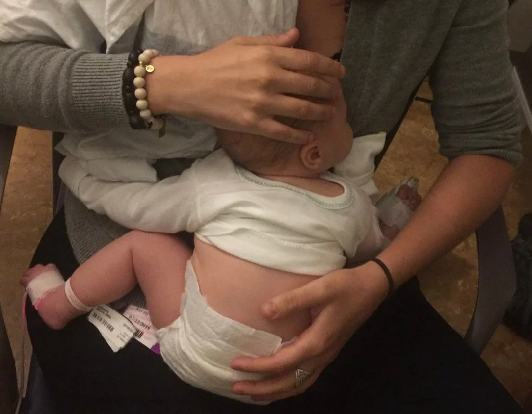 A baby lying on Kyng's lap, receiving a gentle head massage, with the adult supporting the baby's head and holding the baby's body. The baby is wearing a diaper and a white shirt, and has a small medical bandage on one foot.