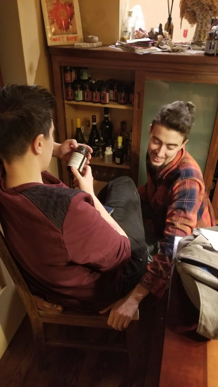 Kyng & Edea sitting close together, one on a wooden chair and the other kneeling, happily interacting in a cozy, warm-lit room. Kyng is holding a small bottle of herbal medicine, and there are various bottles and items on a shelf in front of them.
