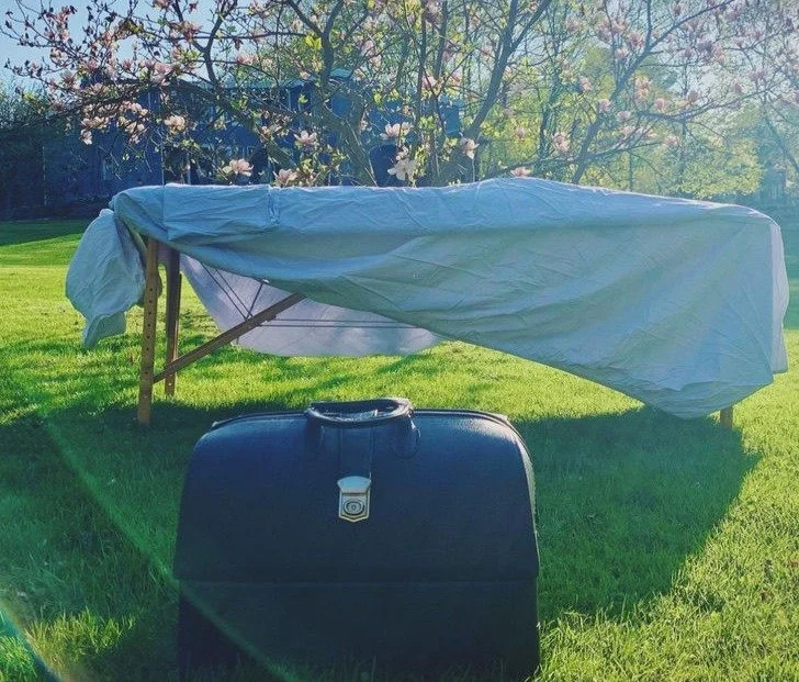 A massage table with a white sheet draped across it in a sunny backyard with green grass, trees, and flowering bushes. In the foreground is a black leather medicine bag.