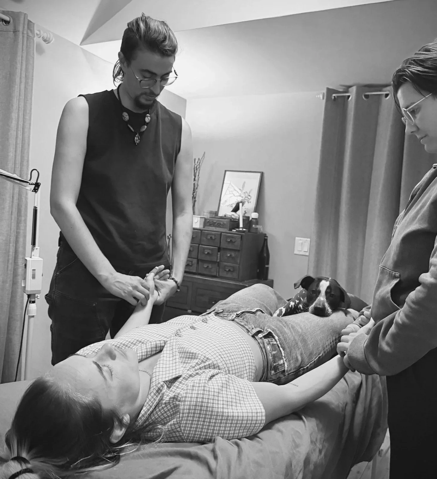 A person lying face-up on a massage bed while receiving 4-hands treatment from both Kyng & Edea at the same time. On their legs Mu, a small dog, provides additional support.