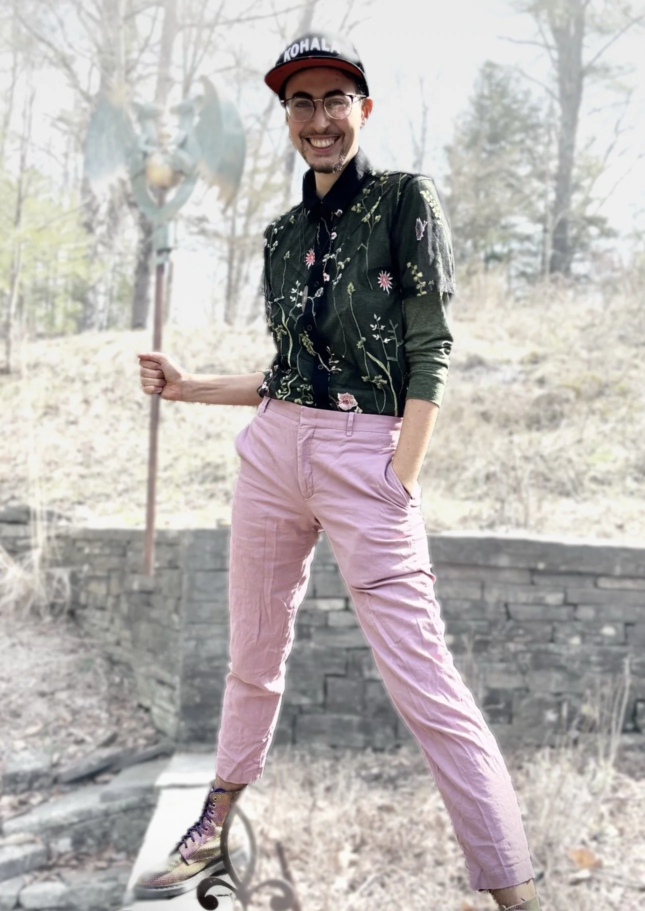 Edea stands outside with a big smile, a staff of twin dragons in one hand, while wearing an outfit with a black embroidered shirt, pink pants, & colorful shoes. They are outdoors on the land at the Center surrounded by trees and grass.