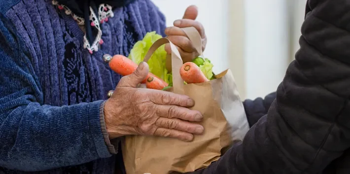 Two people exchanging a paper grocery bag filled with carrots and lettuce.