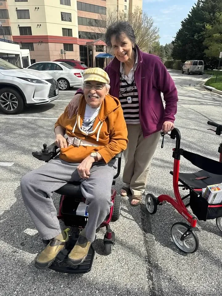 Two women smiling outdoors in a parking lot, one sitting in a mobility scooter and the other standing beside her with a walker.