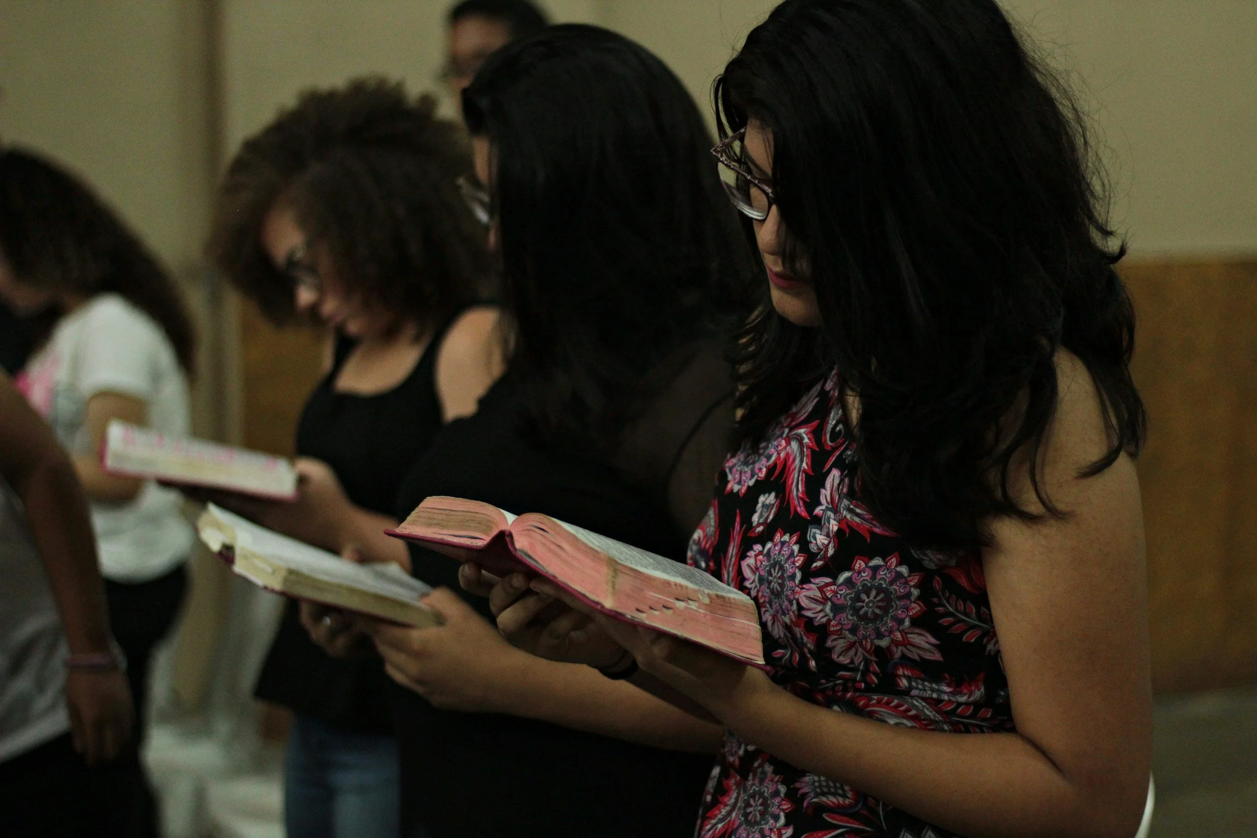 A group of women standing side by side, reading small books in a room with plain walls.
