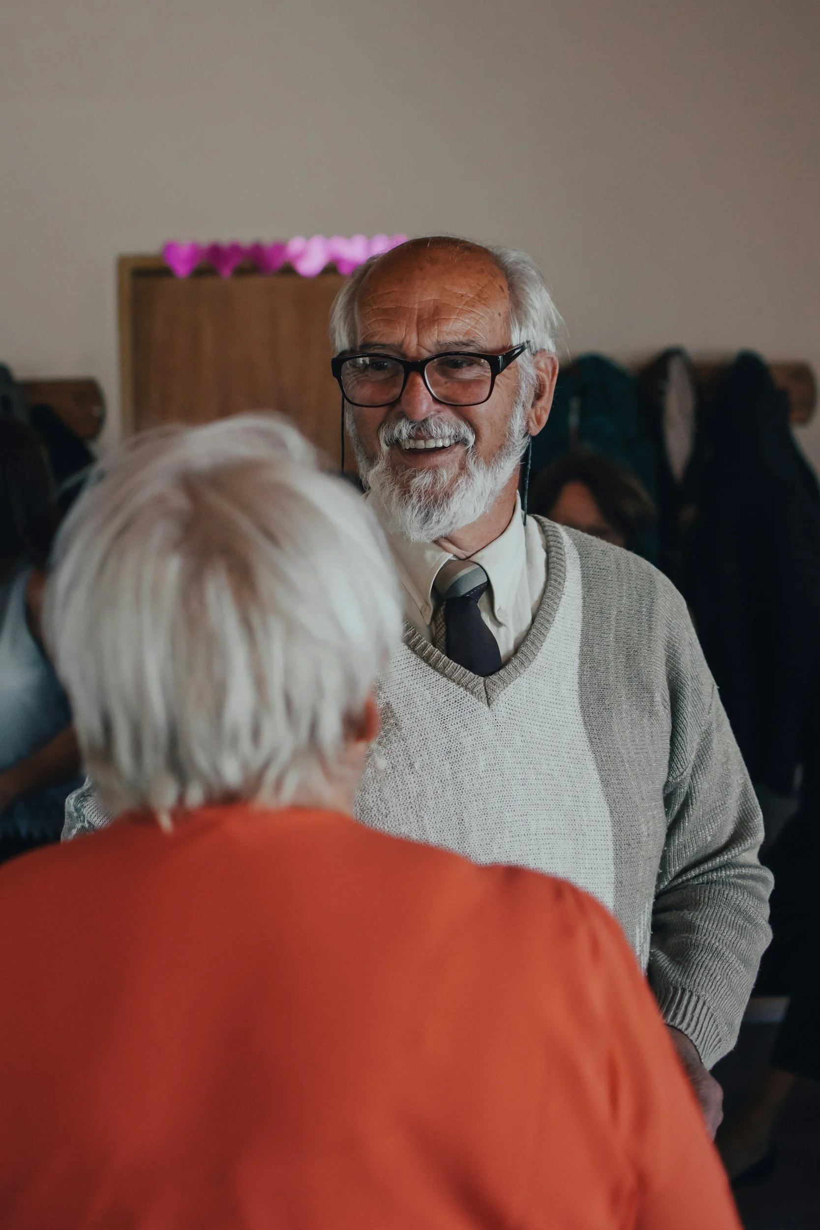 An elderly man with glasses, gray hair, and a beard smiling and engaging in conversation with a woman with white hair in the foreground. The background shows a coat rack and pink flowers.