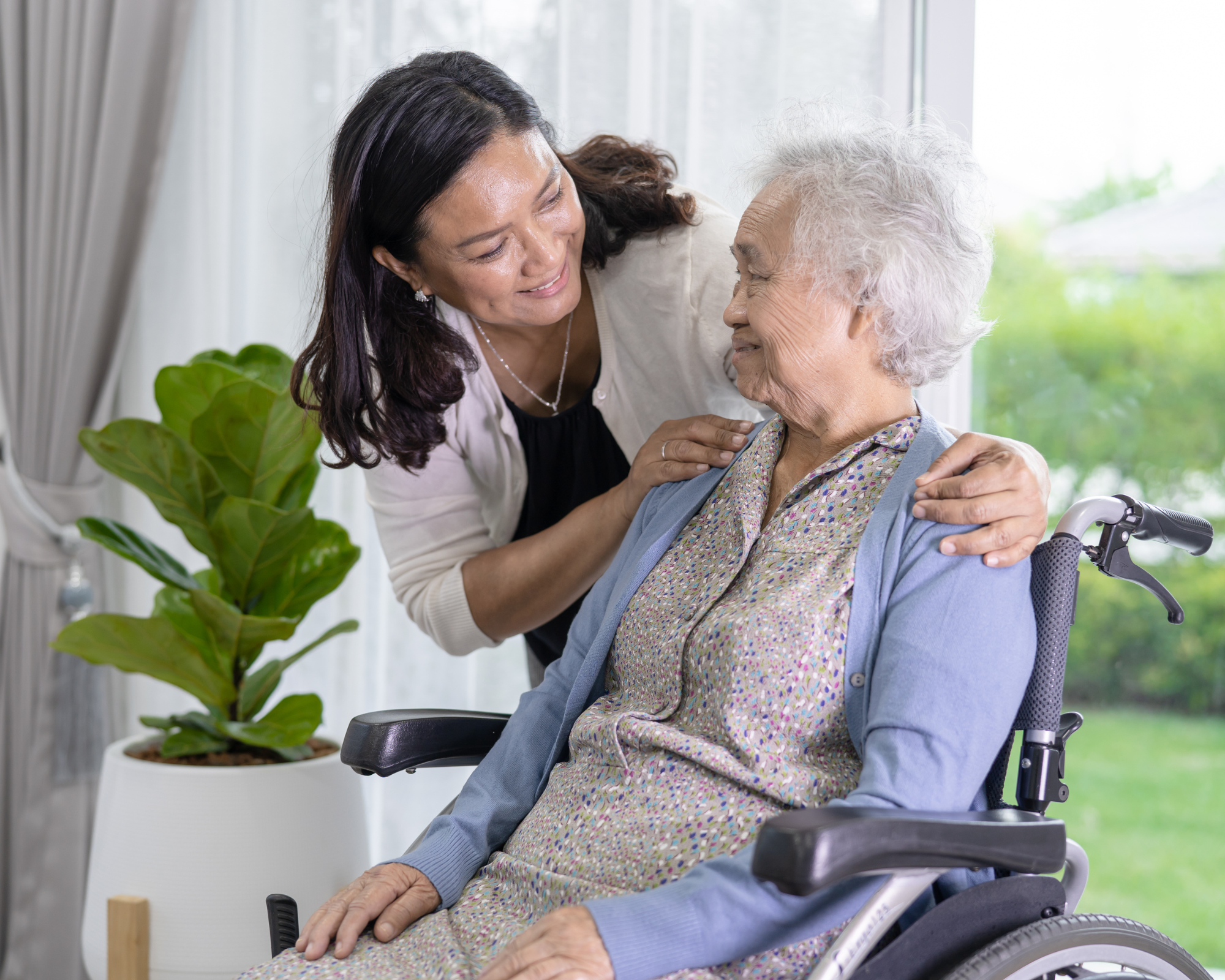 A caregiver smiling and embracing an elderly woman with white curly hair, sitting in a wheelchair, in a bright room with large windows and a green plant nearby.