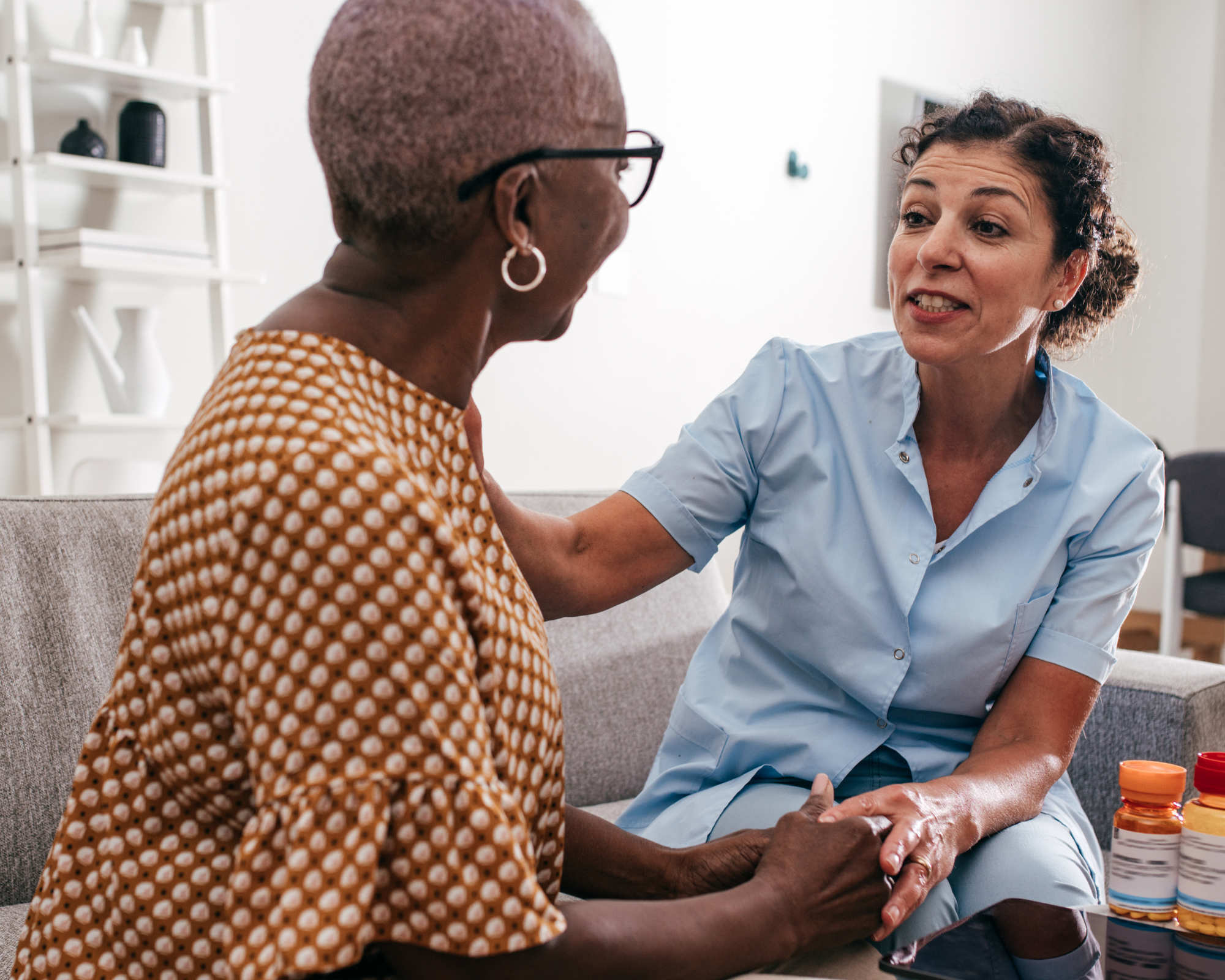 A healthcare worker, wearing a light blue uniform, sitting on a gray couch, holds hands with an elderly woman with short gray hair and glasses, who is wearing a brown patterned dress, in a clinical or home setting with medicine bottles nearby.