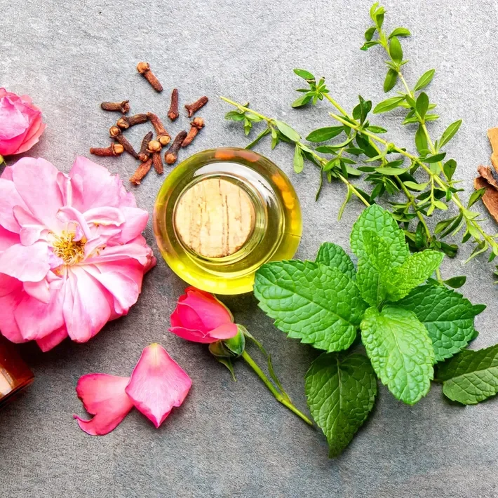 A flat lay of pink roses, green herbs, a small bottle of essential oil, and dried flower petals on a gray surface.