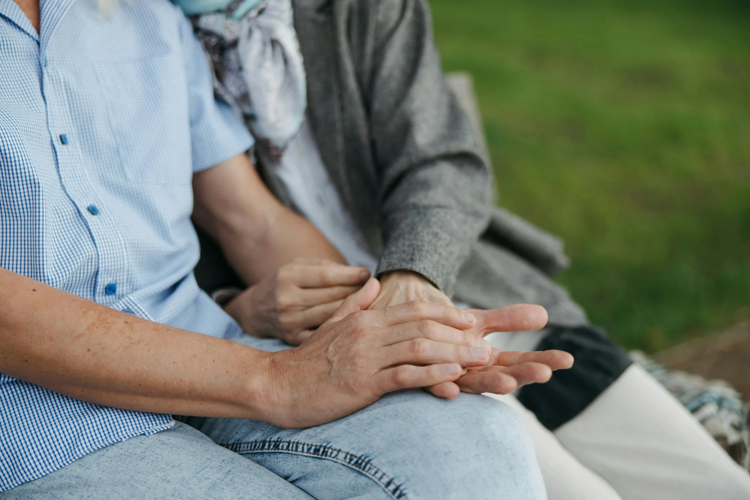 Close-up of two people sitting side by side with their hands folded together, outdoors on a bench with grass in the background.