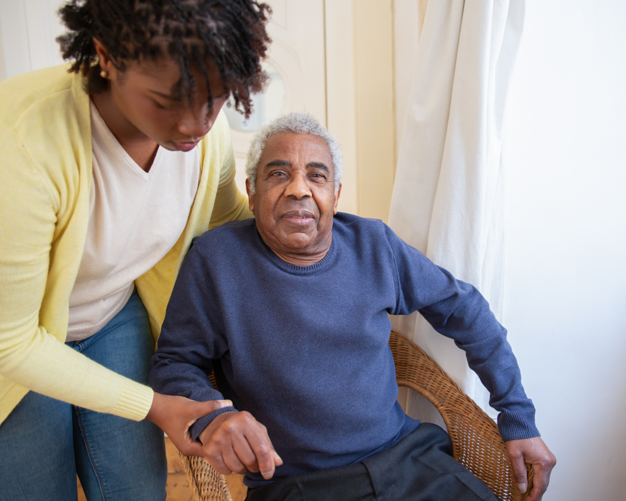 An elderly man with gray hair sitting in a wicker chair, looking at the camera. A woman standing beside him, with her head slightly bowed, gently holding his hand. The woman is wearing a yellow cardigan over a white shirt, and the man is wearing a navy blue sweater. They are indoors near cream-colored curtains and light-colored walls.
