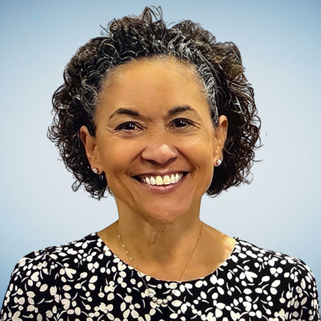 Smiling woman with short, curly dark hair, wearing a black and white patterned top, earrings, and a necklace, against a blue background.