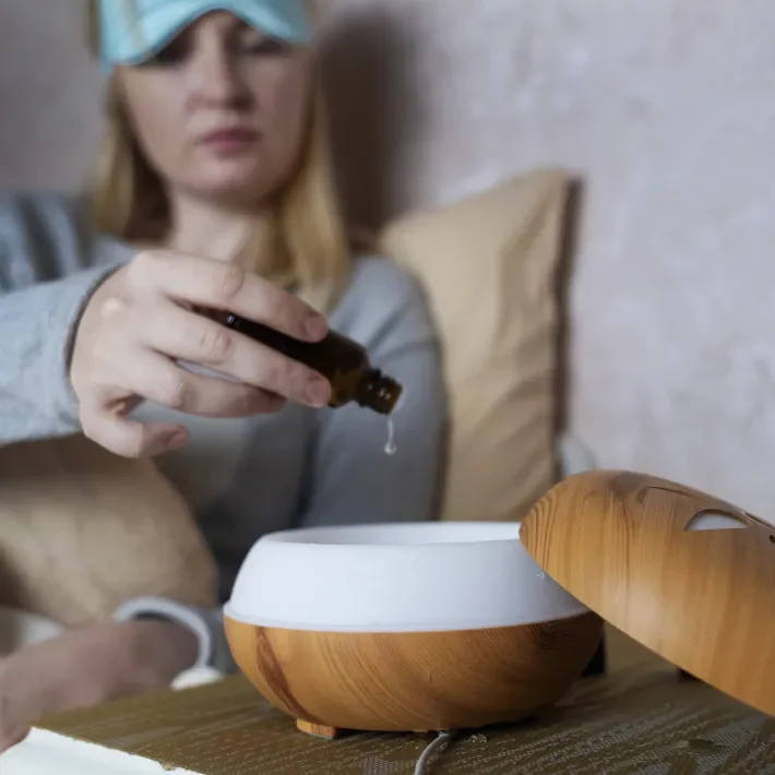 Woman adding essential oil drops into a diffuser on a wooden table.