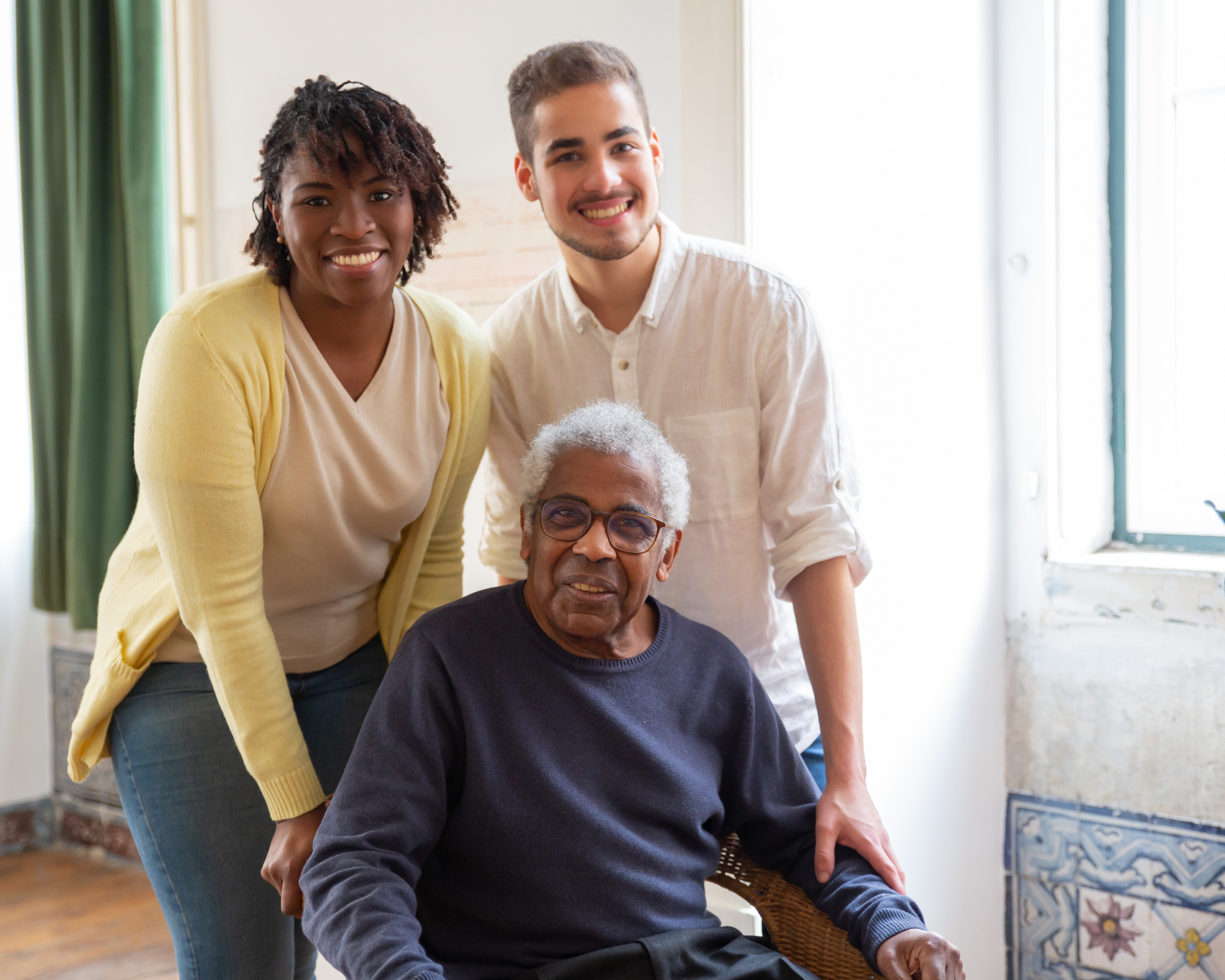 A diverse group of four people, including an elderly man with glasses, a woman with curly hair, and two young men, gathered inside a bright room with large windows and artwork, smiling at the camera.