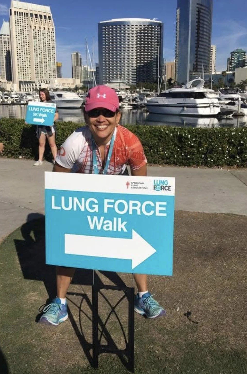 A woman in a pink cap, sunglasses, and athletic clothes kneeling next to a large sign for the Lung Force Walk, pointing to the right. In the background, there are boats docked in a marina and tall buildings under a clear blue sky.
