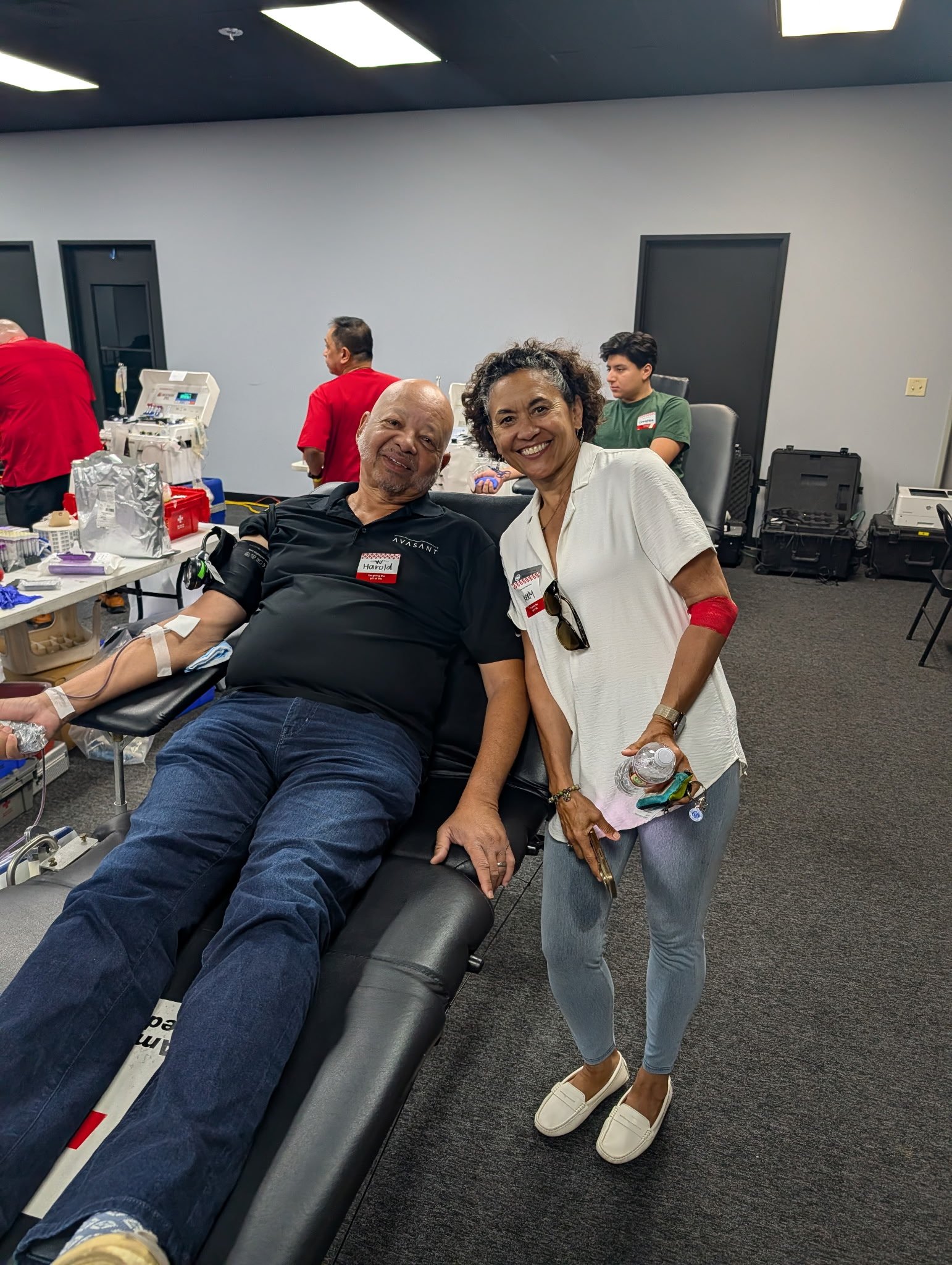 A man and a woman smiling for a photo in a medical donation setting. The man is lying on a reclining chair with medical supplies nearby, and the woman is standing beside him holding a water bottle, both wearing name tags.