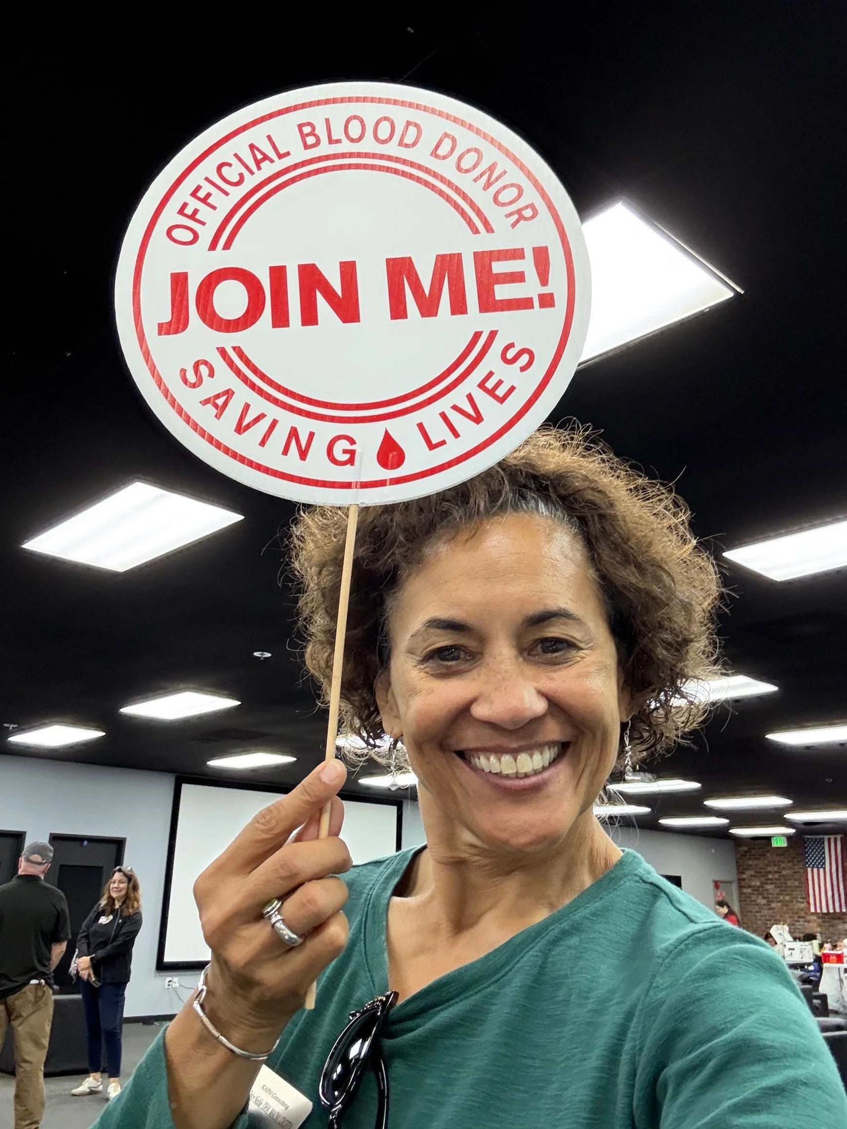 A woman with curly hair smiling and holding a sign that says 'Join Me!' at a blood donation event.