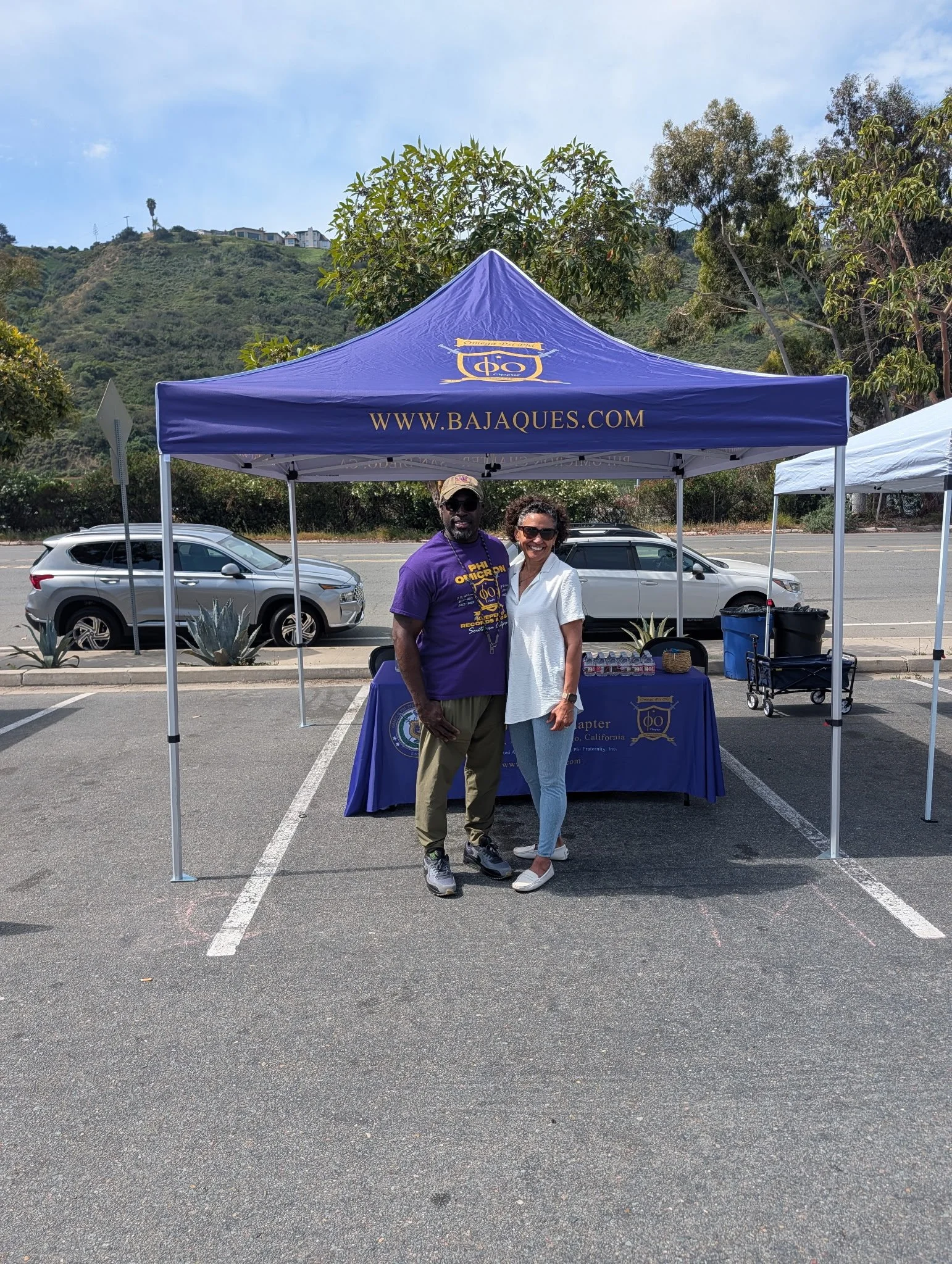 Two smiling people standing under a blue canopy tent on a parking lot, with cars and hills in the background.