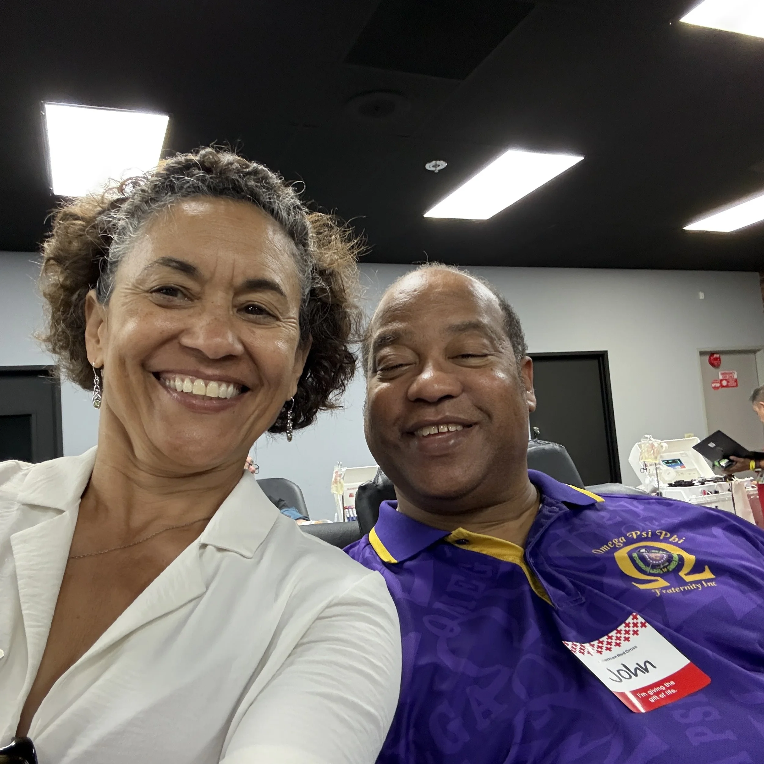 Close-up of two smiling people, a woman with curly hair and a man, wearing a purple shirt with yellow accents, in a room with a black ceiling with fluorescent lights and a gray wall with a door. The woman is holding the camera for a selfie, and the man has a name tag that reads 'John' and is part of a fraternity or organization.