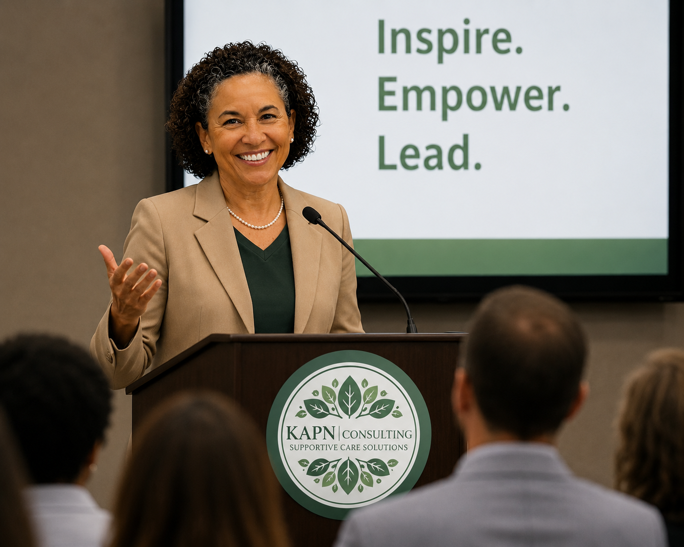 A woman with curly hair, wearing a beige blazer and a pearl necklace, is smiling and speaking at a podium with a microphone in front of an audience. Behind her, a screen displays the words 'Inspire. Empower. Lead.'