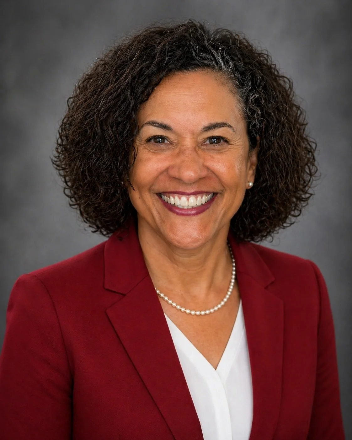 A smiling woman with curly hair wearing a red blazer, white blouse, pearl necklace, and pearl earrings, posing against a gray background.