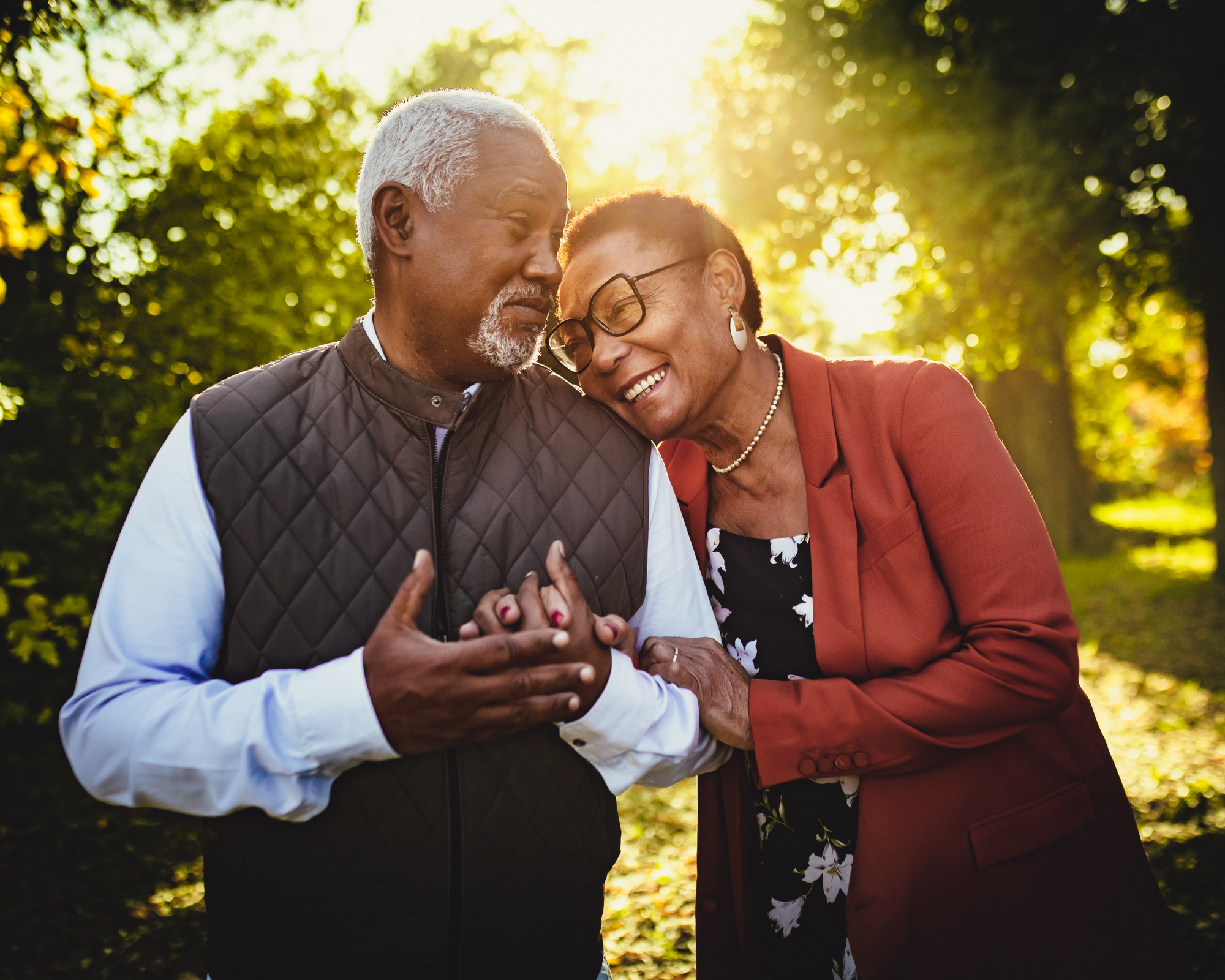 An elderly man and woman sharing a joyful moment outdoors in a park during sunset, their heads touching, smiling and holding hands, surrounded by green trees.