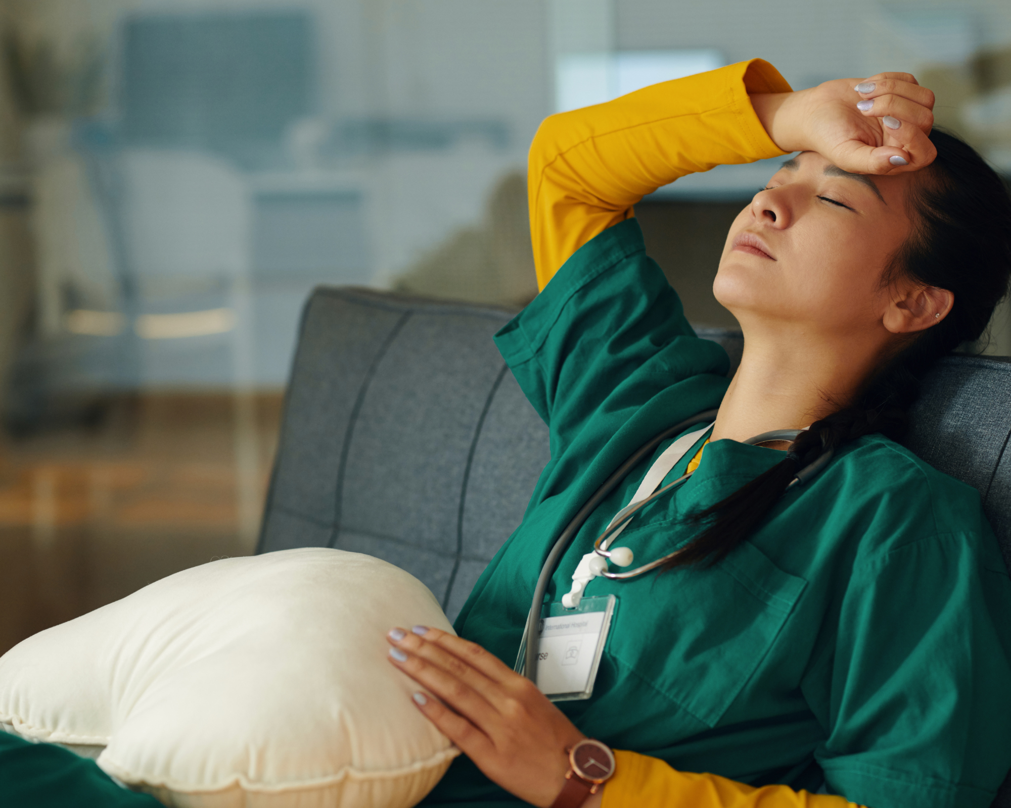 A healthcare worker in green scrubs and a stethoscope resting on a pillow with her hand on her forehead, appearing tired or stressed.