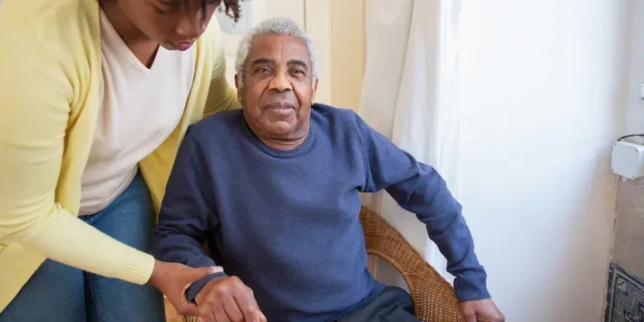 A young person helping an elderly man in a blue sweater, sitting in a wicker chair, inside a room with light-colored curtains.