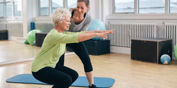 A young female trainer helping an elderly woman with balance exercises at a fitness center.