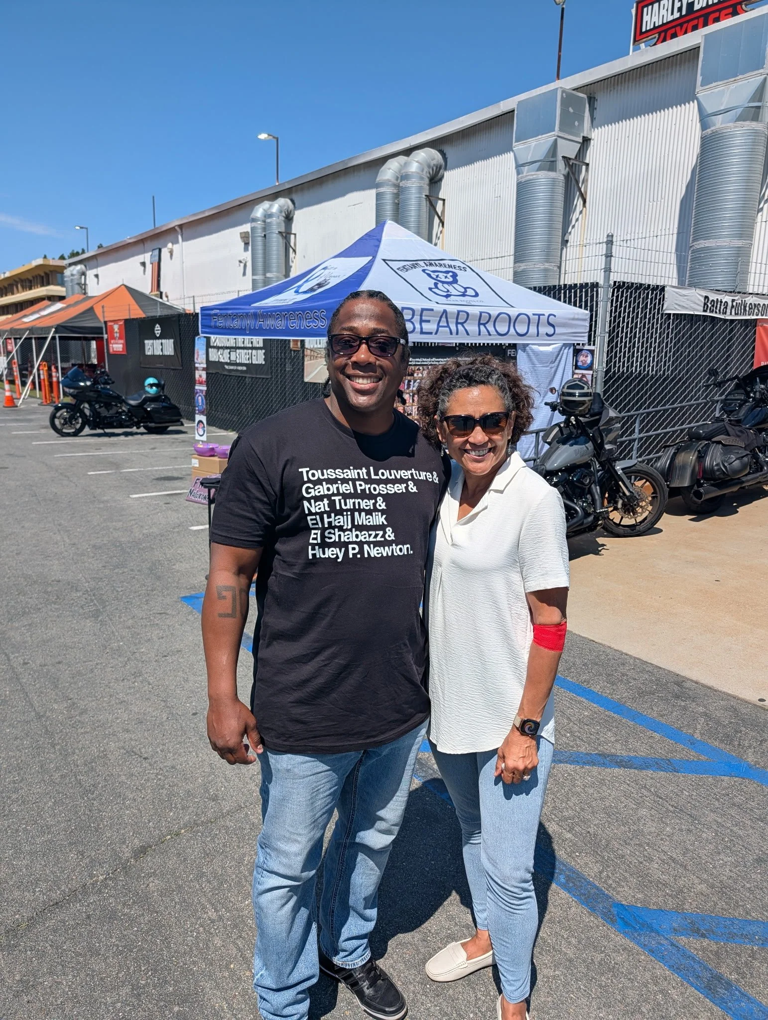 Two smiling people, a man and a woman, standing outside in a parking lot during daytime. The man is wearing a black T-shirt with white text, and the woman is in a white jacket and sunglasses. Behind them, there are motorcycles and a booth with a blue and white tent that has a sign reading 'Fentanyl Awareness' and some text about bear roots. In the background, there's a large industrial building with vent pipes and a parking lot with orange cones.